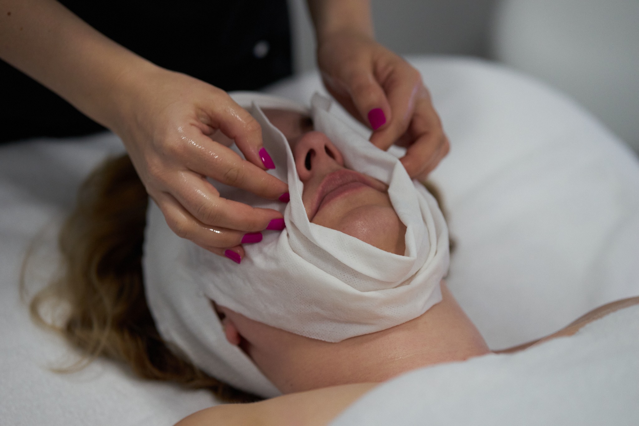 Klientka z nałożoną białą maską na twarzy, dłonie kosmetyczki z różowym manicure poprawiają ułożenie maski