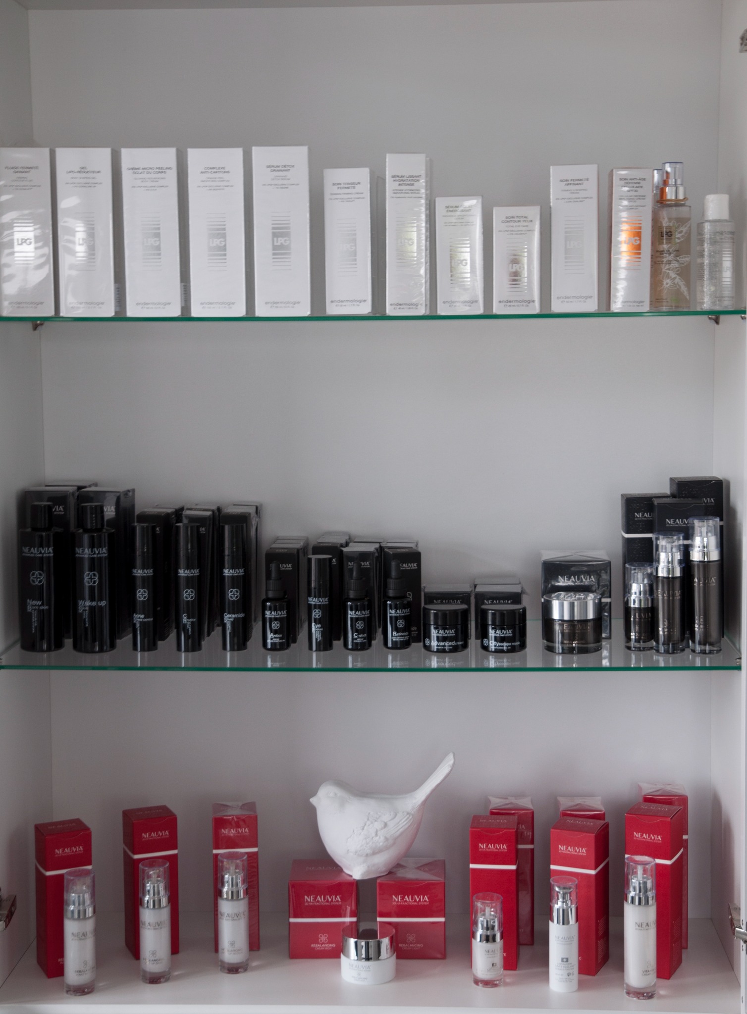 Półka w gabinecie z kosmetykami i preparatami marek LPG i Neauvia, uporządkowane w białych i czarnych opakowaniach, z dekoracyjną białą figurką ptaka.