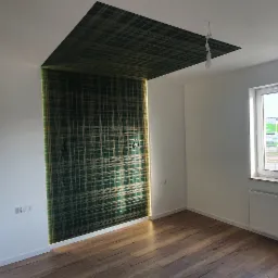 Pokój po remoncie z drewnianą podłogą, białymi ścianami i fragmentem ściany z wzorzystą tapetą w odcieniach zieleni i złota, która przechodzi na sufit tworząc ciekawy efekt wizualny. Widoczne...