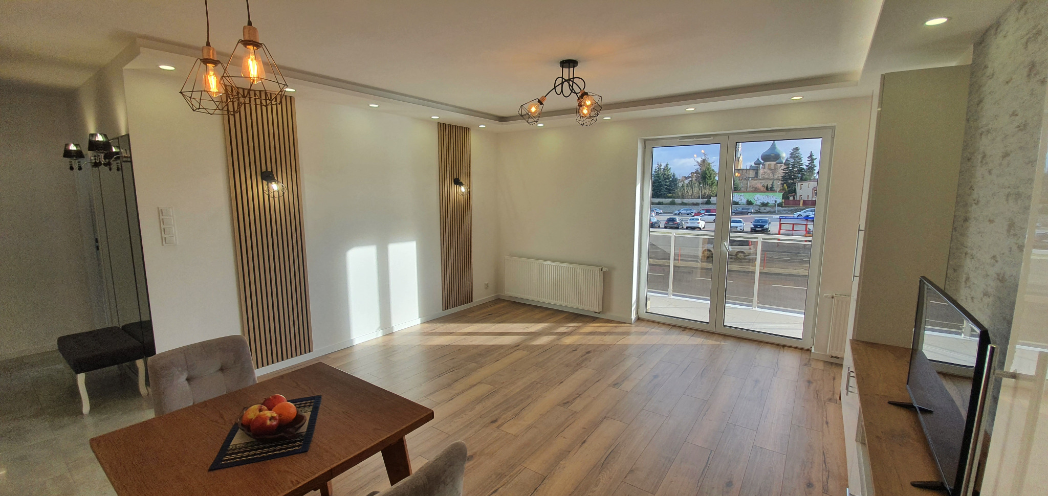 Jasne, przestronne wnętrze po remoncie z drewnianą podłogą, dekoracyjnymi panelami ściennymi, nowoczesnym oświetleniem i widokiem na ulicę z zabytkową cerkwią w tle.