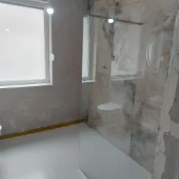 Wnętrze łazienki w trakcie remontu: widoczne marmurowe płytki na ścianach, szklana kabina prysznicowa, biała podłoga z płytek, surowa ściana w stanie deweloperskim, wisząca żarówka tymczasowa.