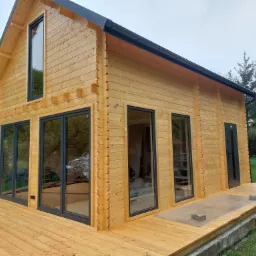 Nowoczesny, parterowy dom z bali w naturalnym odcieniu drewna, z czarnymi ramami okiennymi i drzwiami, otoczony drewnianym tarasem i zielenią.