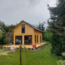 Nowoczesny, drewniany domek letniskowy z czarnymi ramami okiennymi i dachem, posadowiony na betonowych bloczkach, z częściowo ukończonym drewnianym tarasem i skrzynką narzędziową Hilti na nim...
