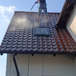 Pracownik w uprzęży zabezpieczającej czyści dach z dachówki ceramicznej za pomocą myjki ciśnieniowej; widoczny świetlik dachowy i rynna.