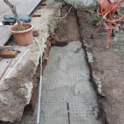 Wykop pod fundament z siatką zbrojeniową zalany świeżym betonem, widoczna pomarańczowa betoniarka, donica z uschniętym drzewkiem oraz fragment drewnianego tarasu w tle.