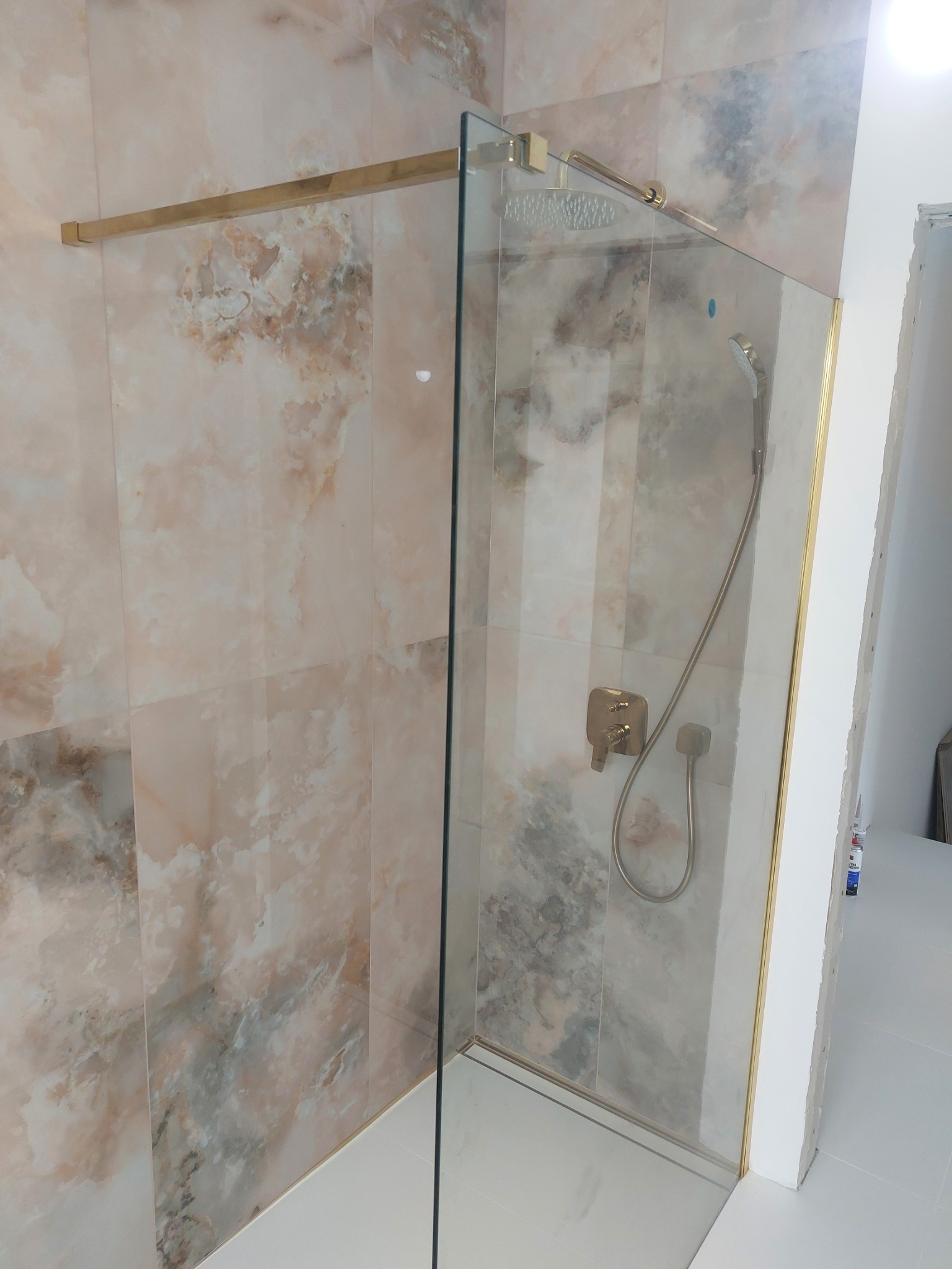 Kabina prysznicowa typu walk-in z armaturą w kolorze złotym, osadzona na białych płytkach podłogowych, z tłem z płytek imitujących marmur.
