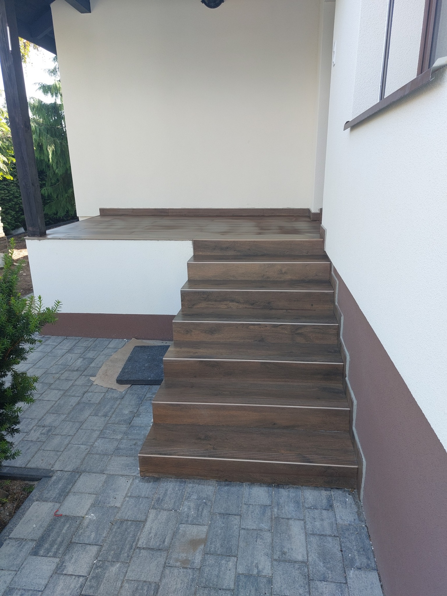 Zewnętrzne schody wejściowe wyłożone płytkami imitującymi drewno, z szarymi fugami i metalowymi listwami antypoślizgowymi, prowadzące na taras przed domem, z szarą kostką brukową na podłożu.