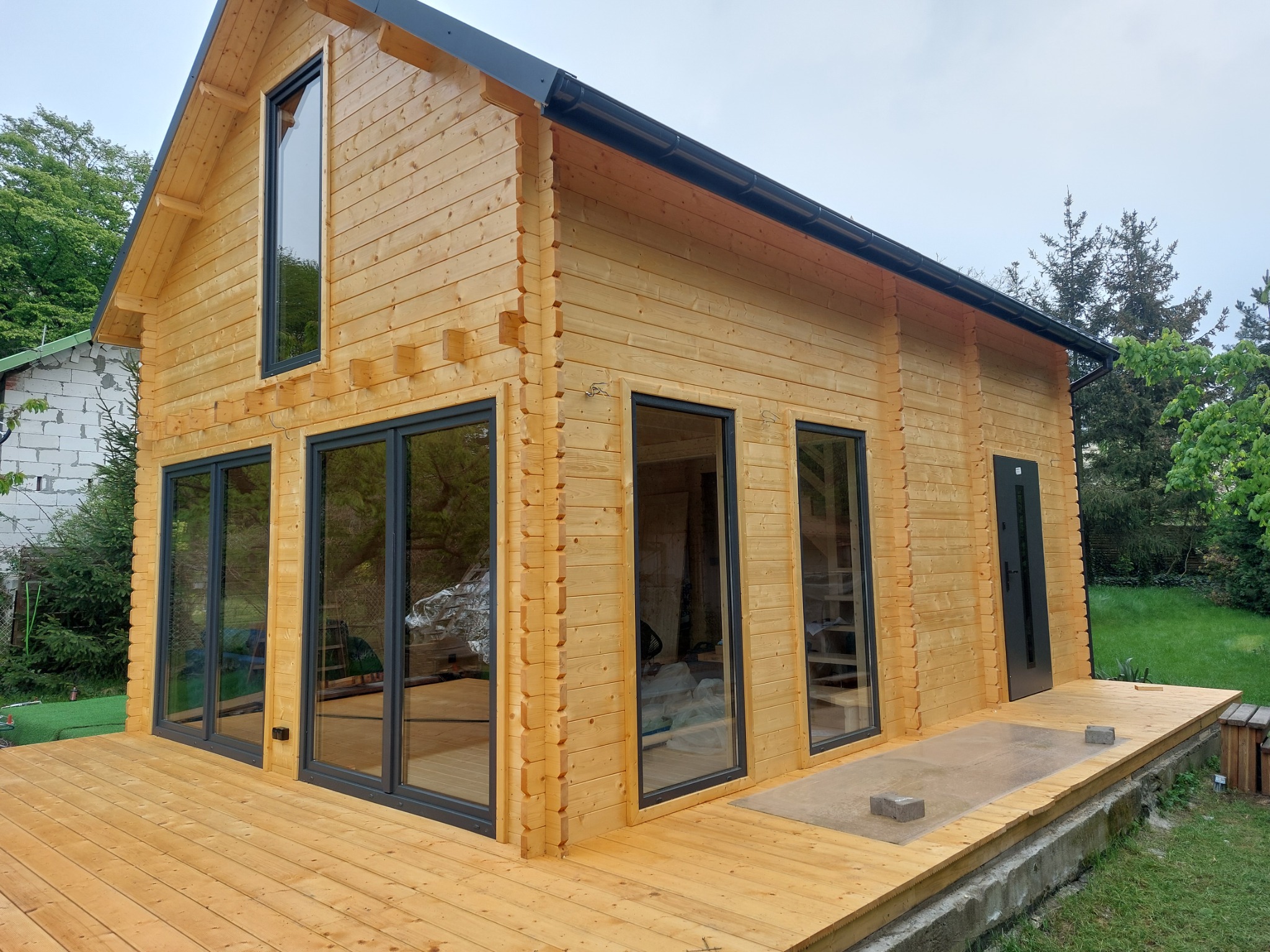 Nowoczesny, parterowy dom z bali w naturalnym odcieniu drewna, z czarnymi ramami okiennymi i drzwiami, otoczony drewnianym tarasem i zielenią.