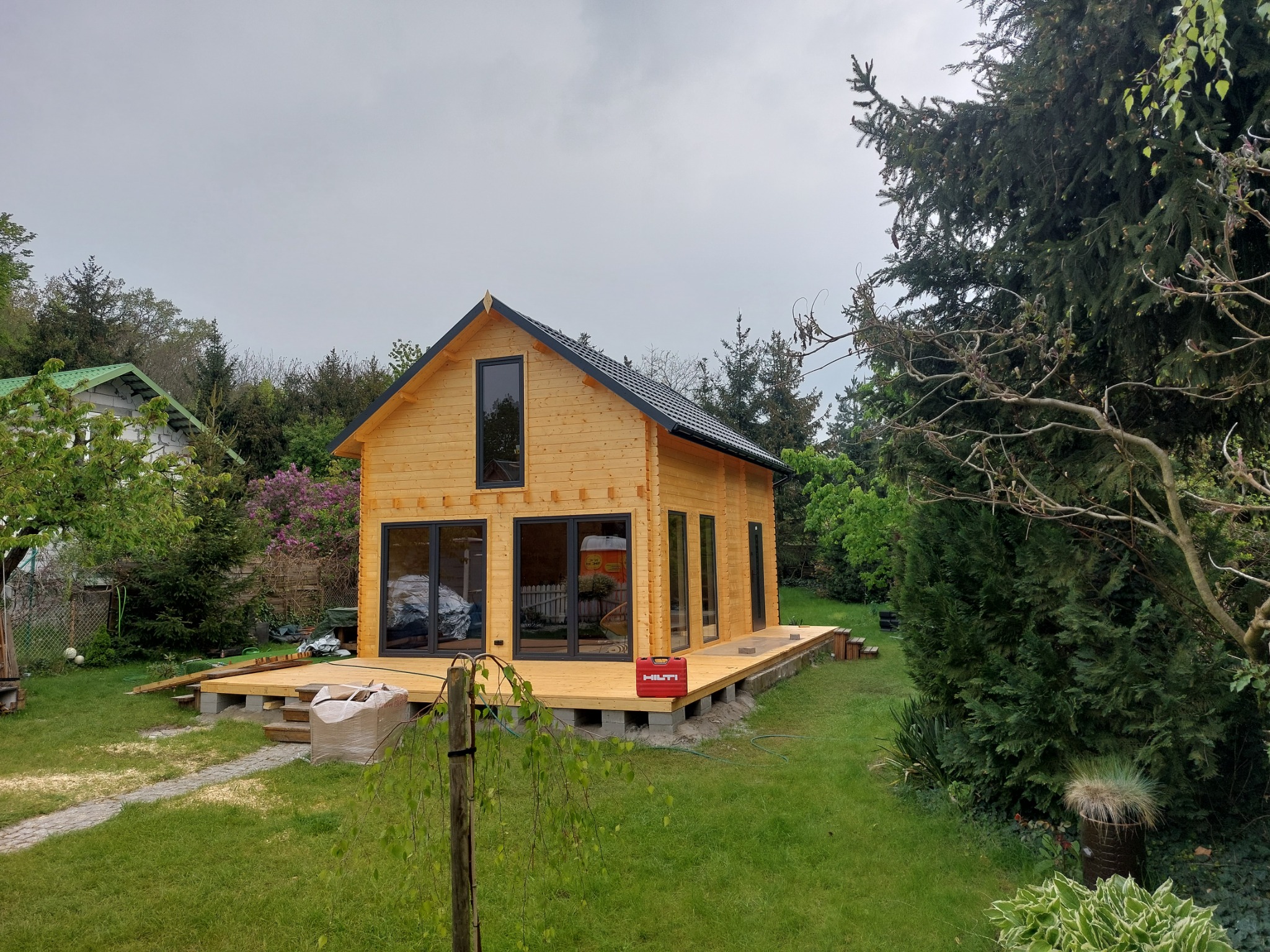 Nowoczesny, drewniany domek letniskowy z czarnymi ramami okiennymi i dachem, posadowiony na betonowych bloczkach, z częściowo ukończonym drewnianym tarasem i skrzynką narzędziową Hilti na nim...