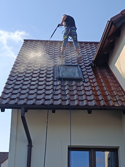 Pracownik w uprzęży zabezpieczającej czyści dach z dachówki ceramicznej za pomocą myjki ciśnieniowej; widoczny świetlik dachowy i rynna.
