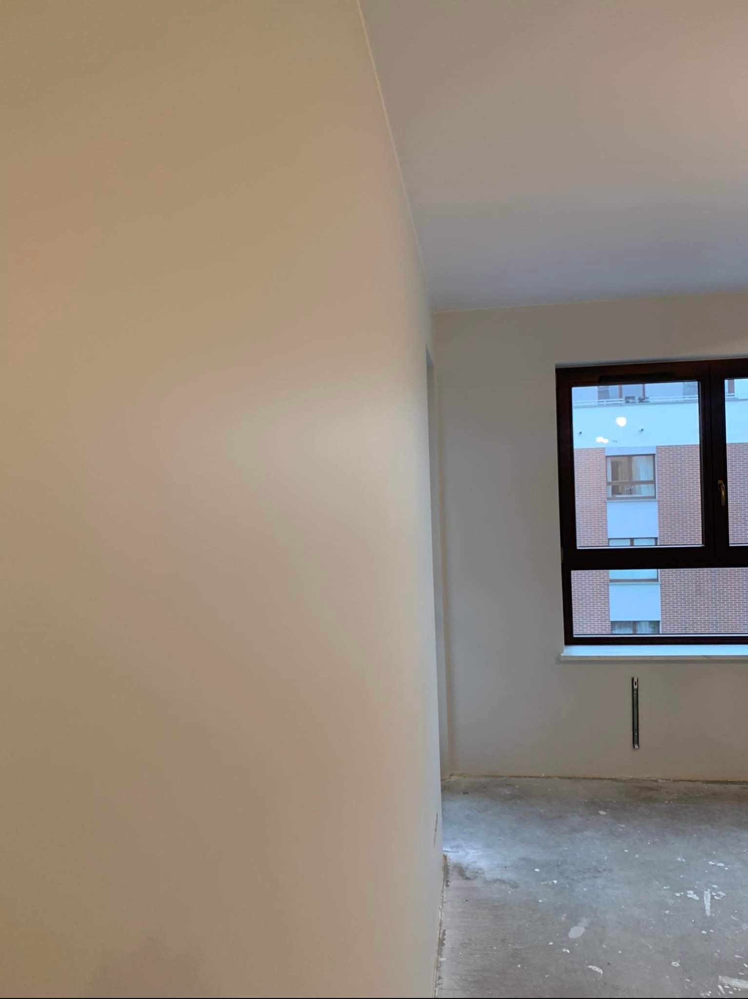 Świeżo pomalowane na beżowo ściany w pustym pokoju z widokiem na budynek z cegły przez ciemnobrązowe okno; podłoga w stanie surowym.