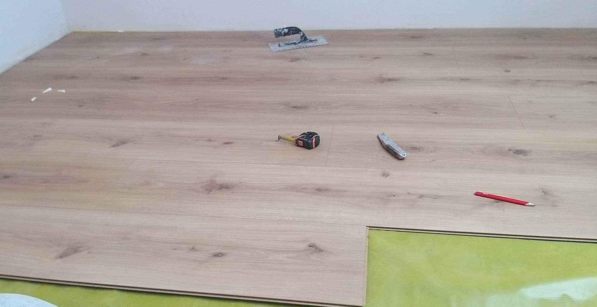 Układanie paneli podłogowych: widoczna podkładka, deski z jasnego drewna, miara, nóż, ołówek i kielnia do kleju.