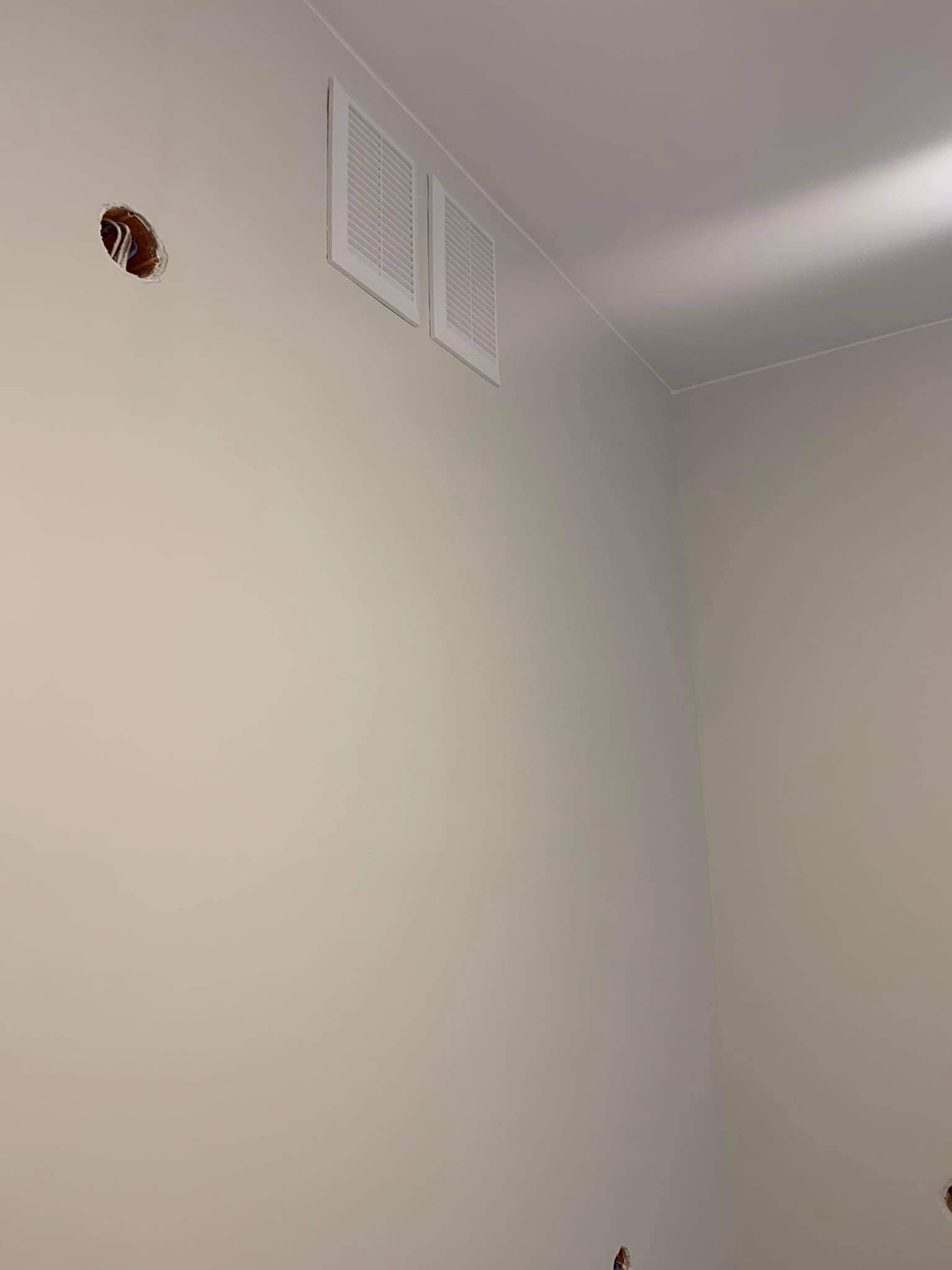 Świeżo pomalowana ściana w jasnym odcieniu z dwoma białymi kratkami wentylacyjnymi i otworem na kabel elektryczny bez wykończenia.