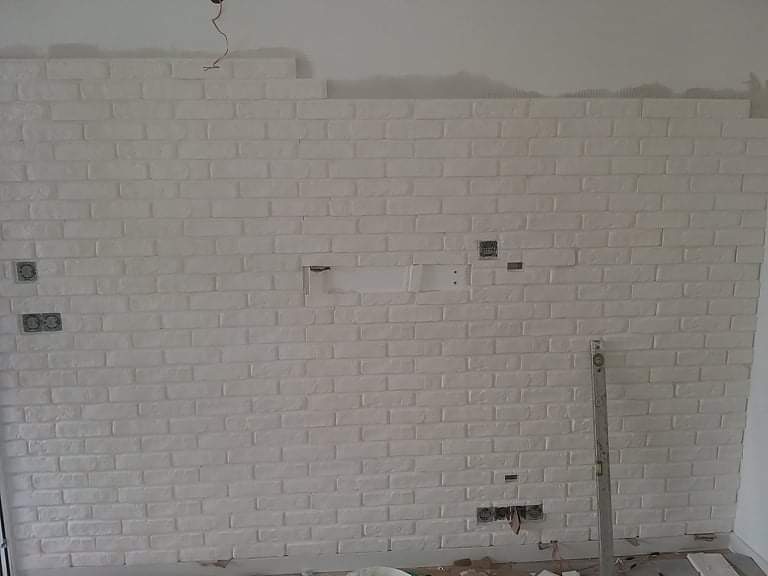 Biała ściana z cegły dekoracyjnej w trakcie montażu, widoczne puszki elektryczne i poziomica przy ścianie.