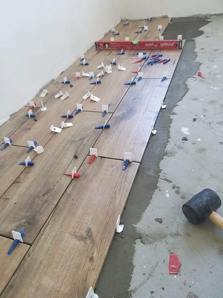 Układanie drewnopodobnych płytek podłogowych z użyciem systemu poziomowania; widoczna poziomica, gumowy młotek i zaprawa.