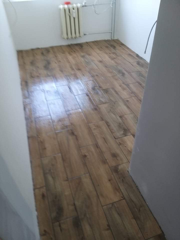 Ułożone brązowe płytki imitujące drewno na podłodze w łazience, widoczny biały grzejnik i rury przy ścianie.