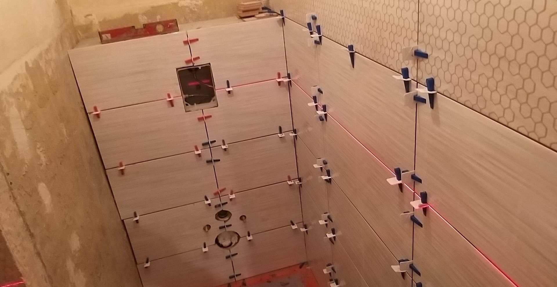 Układanie beżowych płytek w łazience z użyciem poziomicy laserowej i krzyżyków dystansowych, widoczne otwory na instalacje i surowa ściana w tle.