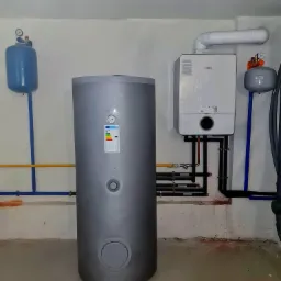 Szary zasobnik ciepłej wody użytkowej z etykietą energetyczną, wiszący biały piec gazowy Bosch z podłączeniami, niebieskie i żółte rury instalacyjne na tle surowej, białej ściany.