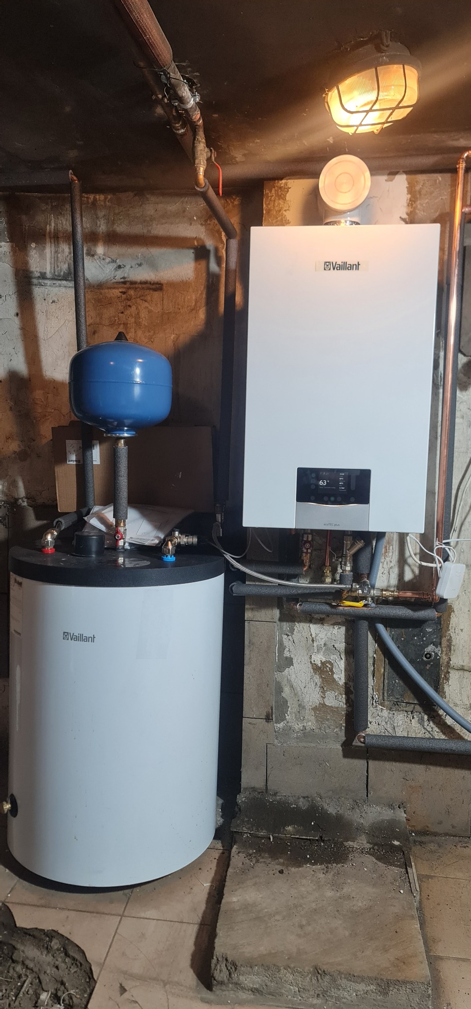 Biały kondensacyjny kocioł gazowy marki Vaillant zamontowany obok zbiornika na wodę w piwnicy z odsłoniętymi rurami i starą lampą na suficie.