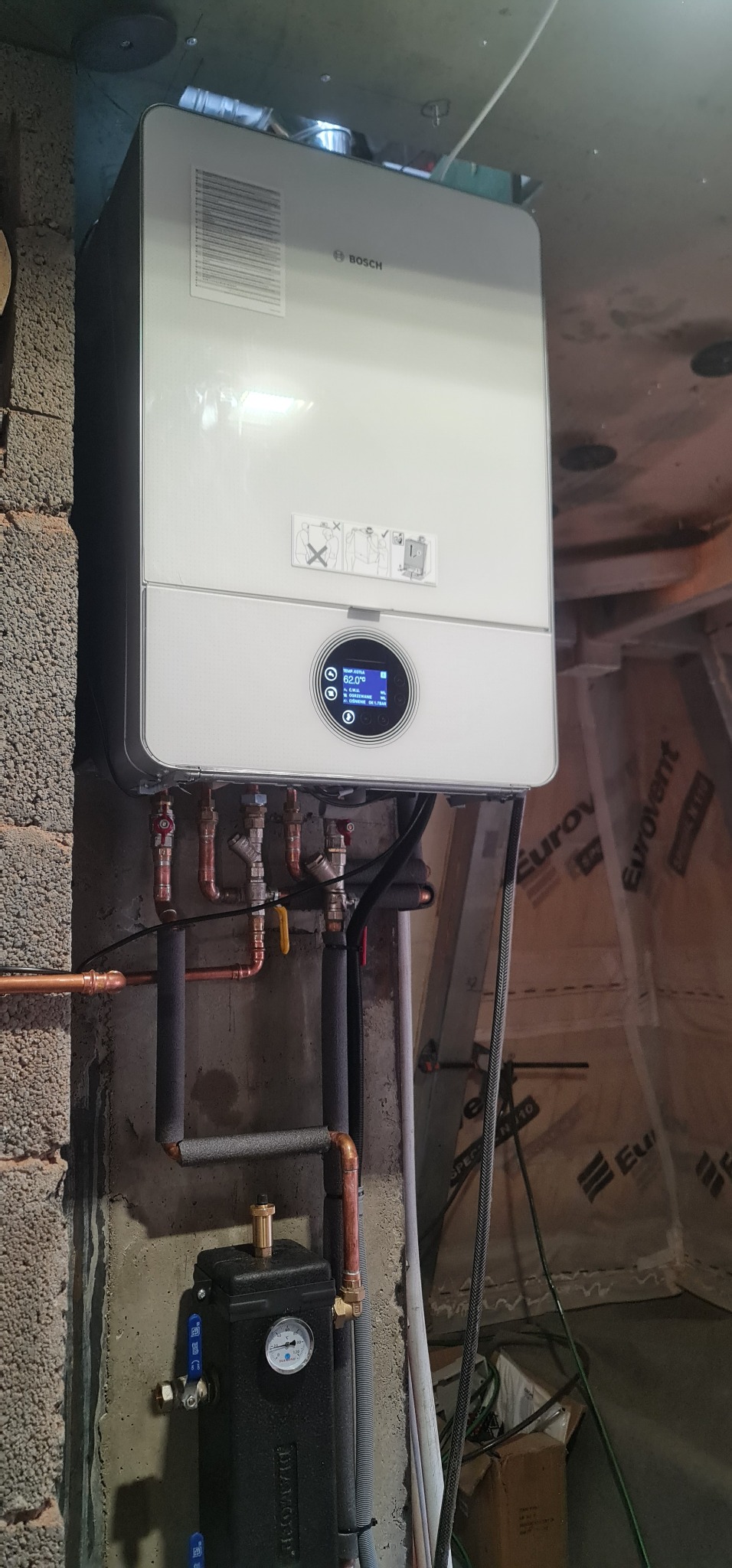 Nowoczesny, wiszący piec gazowy marki Bosch z podłączonymi miedzianymi rurami i zaworami, zamontowany na szarej, betonowej ścianie w surowym pomieszczeniu.