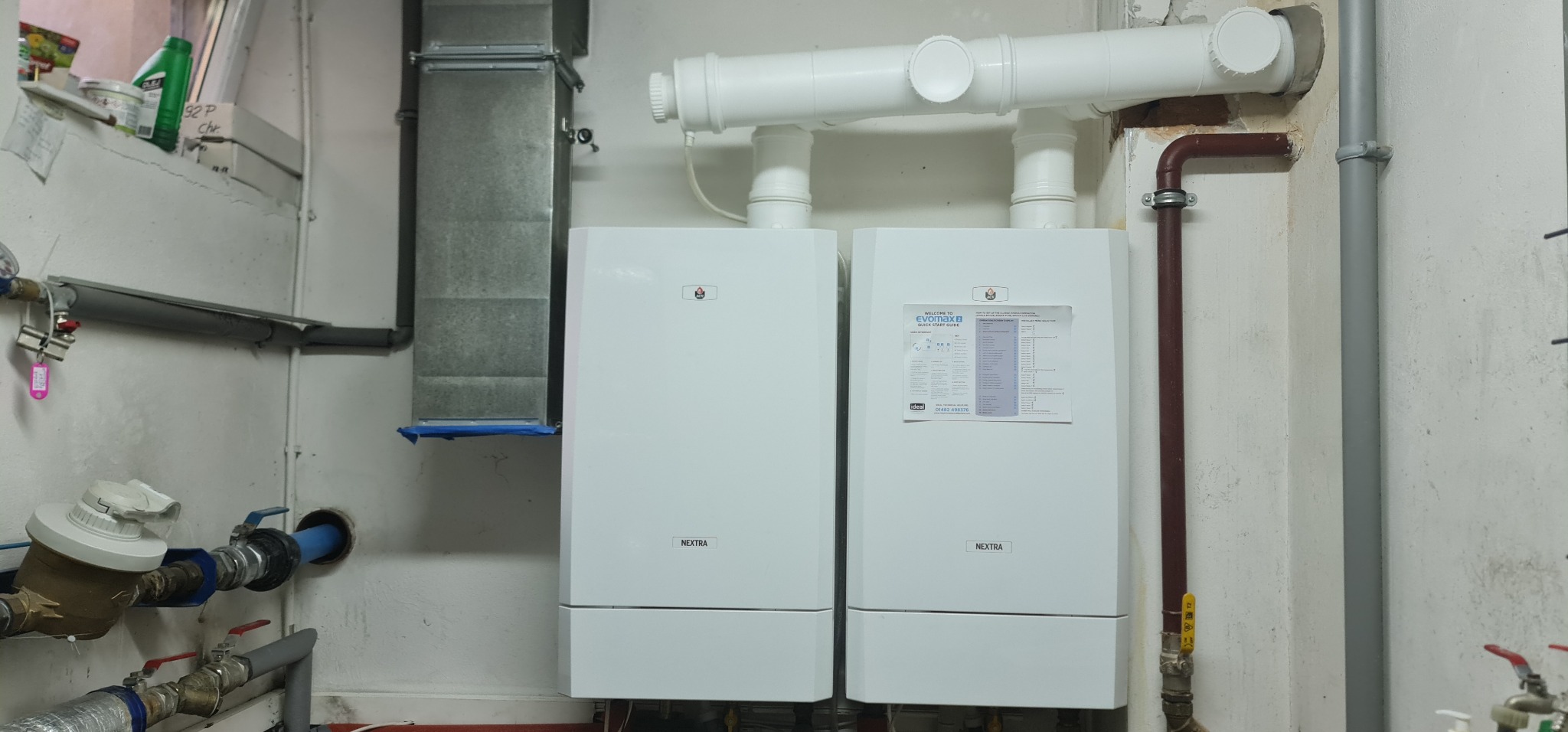 Dwa białe kotły gazowe Nextra zamontowane obok siebie z podłączonymi rurami, widoczne elementy instalacji hydraulicznej i wentylacyjnej w pomieszczeniu o surowym wykończeniu.