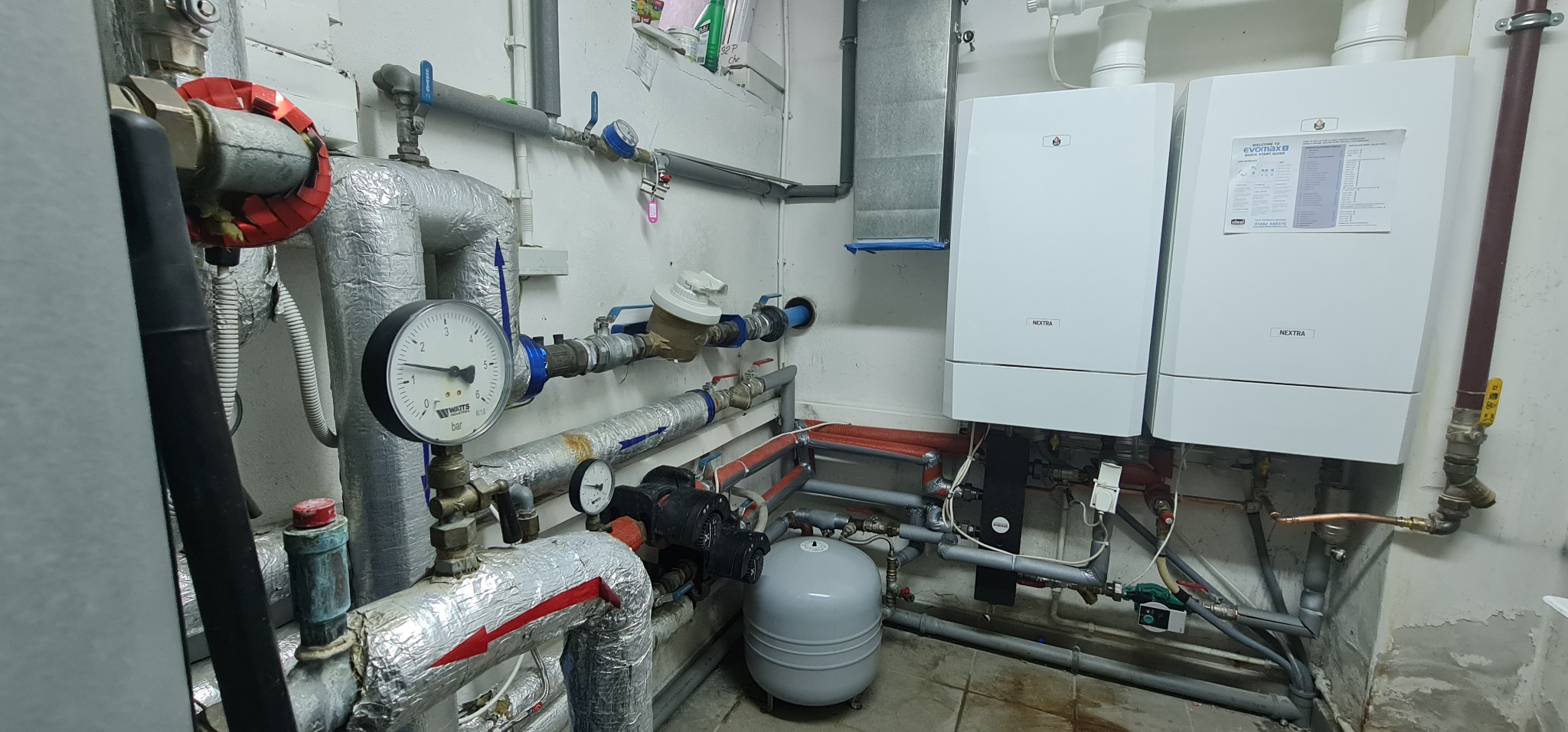 Komora z dwoma białymi kotłami grzewczymi Nextra, rurami izolowanymi folią aluminiową, manometrem Watts i szarym zbiornikiem wyrównawczym, widoczne liczniki i zawory.