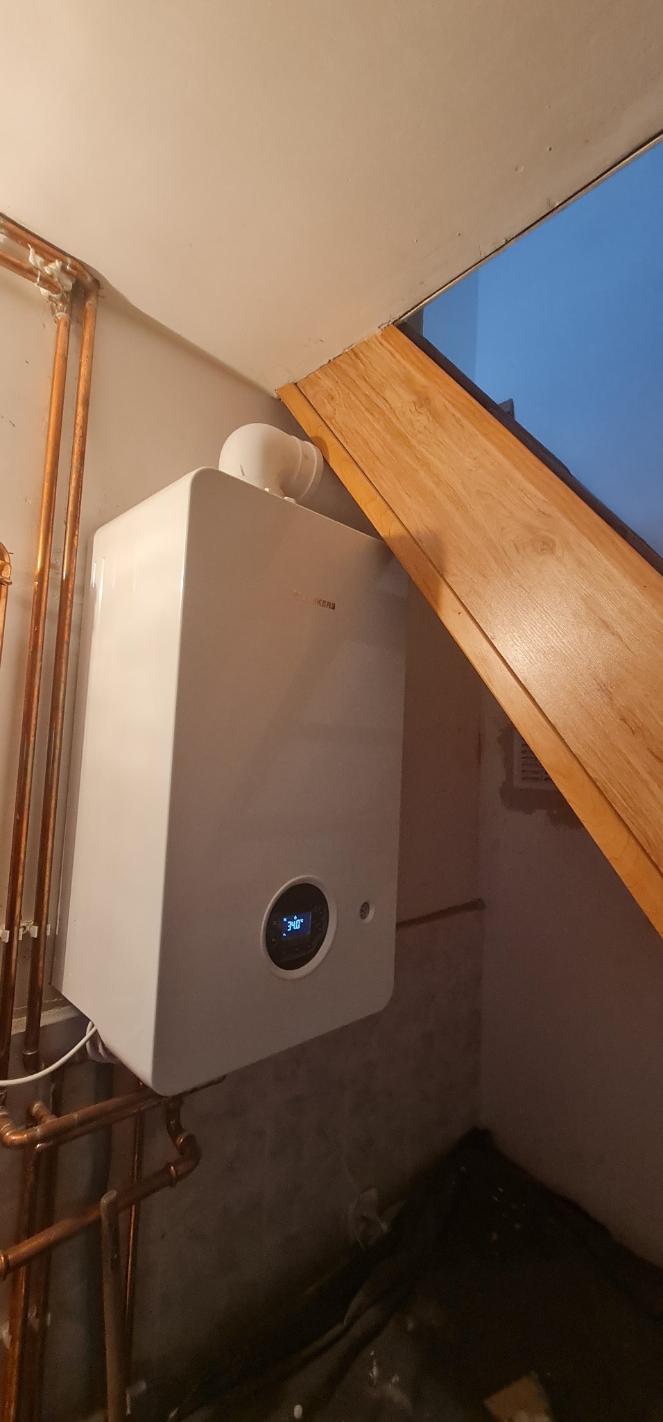 Nowoczesny, biały piec gazowy wiszący na ścianie, z widocznymi miedzianymi rurami instalacji i fragmentem drewnianej konstrukcji skośnego sufitu.