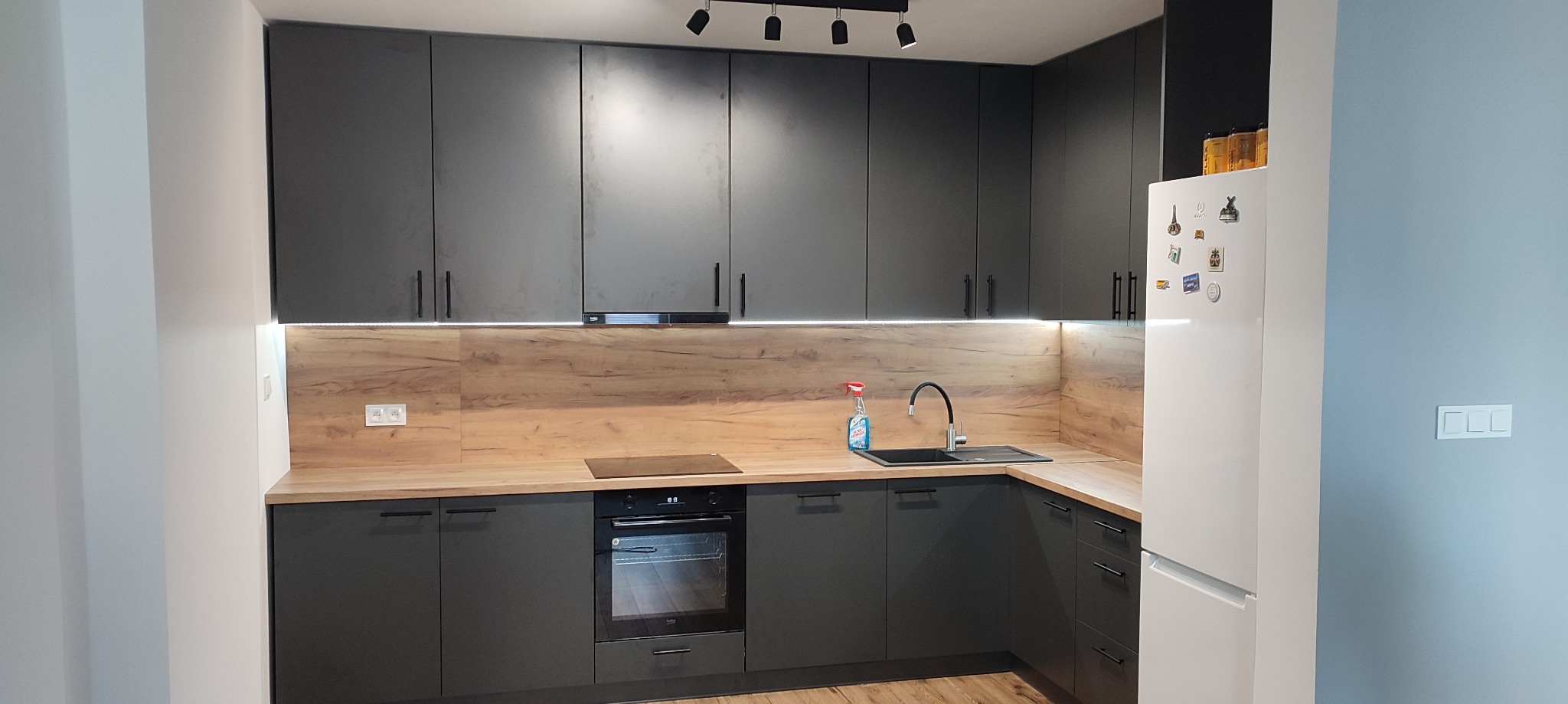 Nowoczesna kuchnia w odcieniach szarości z drewnianym blatem i oświetleniem LED podszafkowym, widoczny czarny zlewozmywak i bateria, biała lodówka z magnesami po prawej stronie.