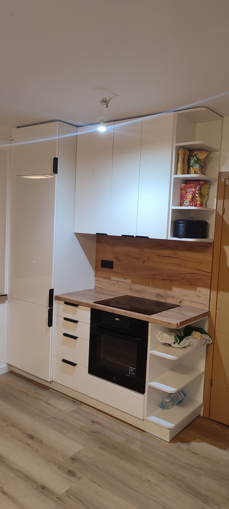 Biała zabudowa kuchenna z drewnianym blatem i panelem, widoczna płyta indukcyjna, piekarnik, szafki z czarnymi uchwytami oraz otwarte półki z artykułami spożywczymi i butelką wody.