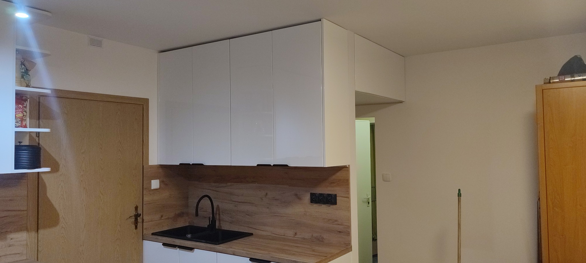 Nowoczesna zabudowa kuchenna z białymi, lakierowanymi szafkami wiszącymi i blatem imitującym drewno, czarny zlew jednokomorowy z baterią, widok na drzwi po lewej stronie i fragment szafy po prawej.