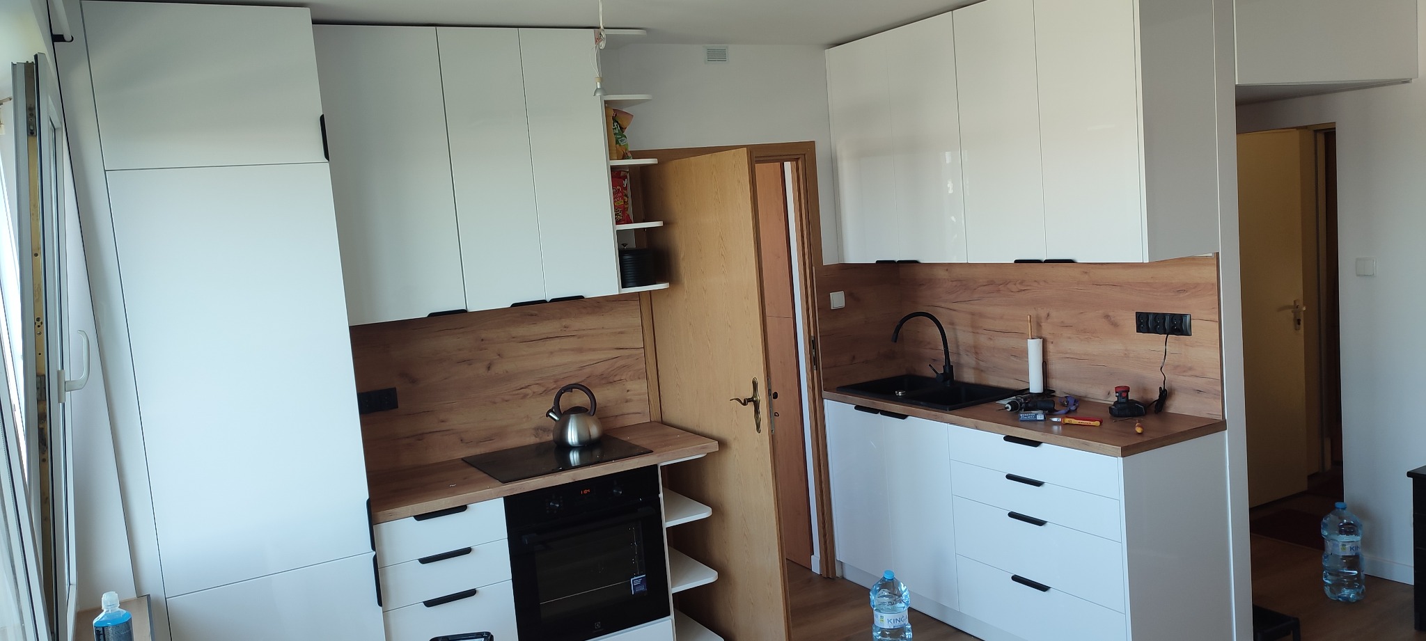 Biała kuchnia z drewnianym blatem i czarnym zlewem, widoczny czajnik na płycie indukcyjnej i butelka wody na podłodze.