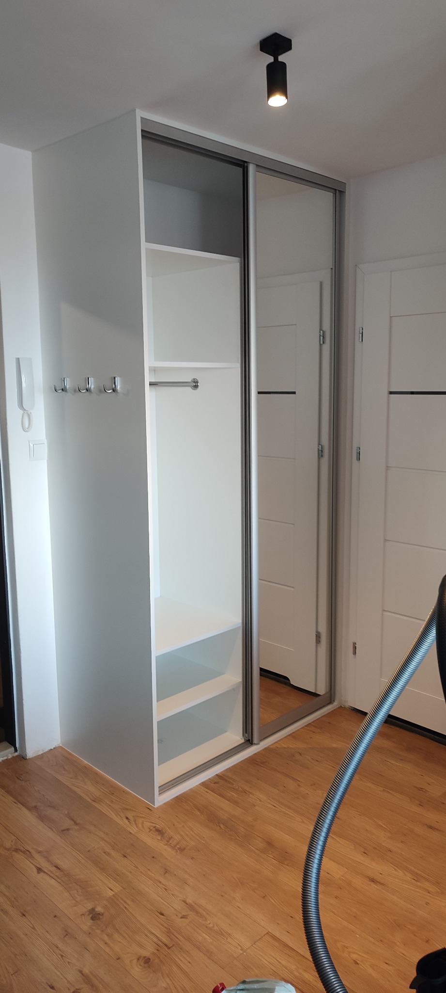 Biała szafa wnękowa z przesuwnymi lustrzanymi drzwiami, widoczne półki i drążek na ubrania, zamontowana w przedpokoju z drewnianą podłogą i wieszakami na ścianie, w tle białe drzwi.
