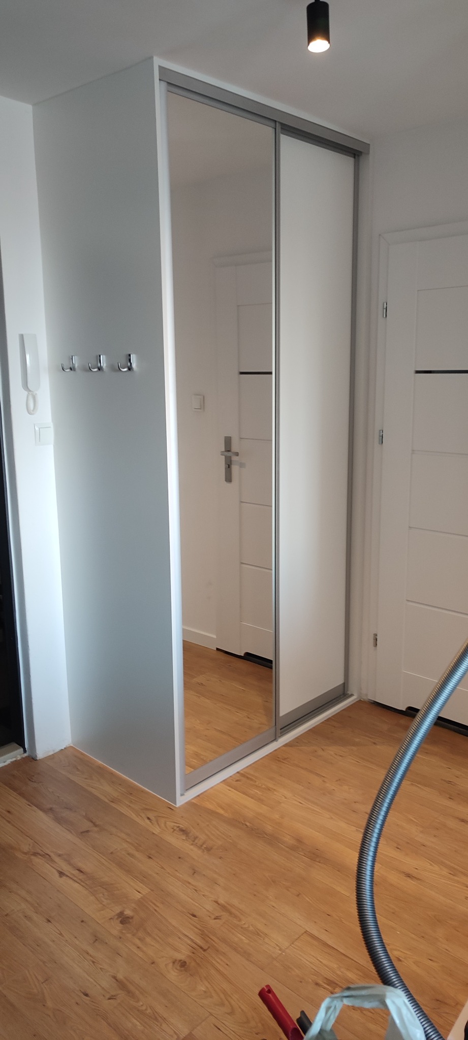 Biała szafa wnękowa z przesuwnymi drzwiami lustrzanymi w przedpokoju z drewnianą podłogą, widoczne wieszaki na ścianie i rura od odkurzacza na podłodze.