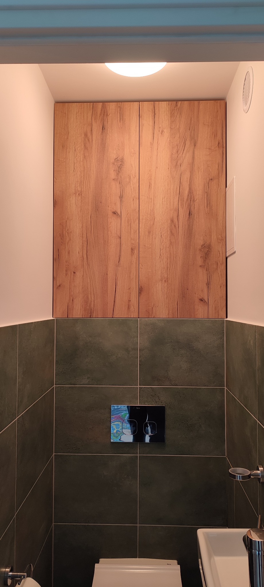 Drewniane szafki nad toaletą w łazience w Warszawie, z ciemnozielonymi płytkami i chromowanym przyciskiem spłukiwania.