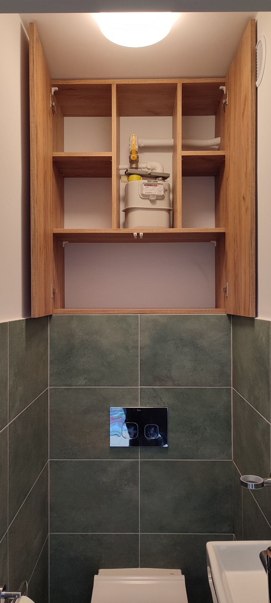 Szafka wisząca w łazience z drewnianymi frontami, zabudowująca licznik gazu, na tle zielonych płytek i białej ceramiki sanitarnej.