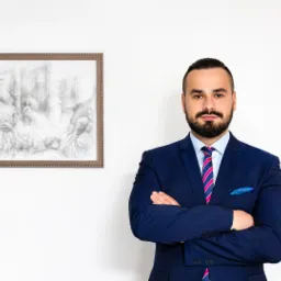 Portret mężczyzny z brodą w niebieskim garniturze, skrzyżowane ręce, w tle na białej ścianie wisi oprawiony rysunek przedstawiający scenę zbiorową z postaciami.