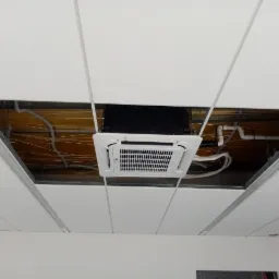 Biała jednostka wewnętrzna klimatyzacji kasetonowej zamontowana w suficie podwieszanym, widoczne rury i przewody instalacyjne w przestrzeni nad sufitem.