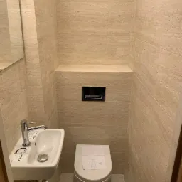 Wąska toaleta wykończona trawertynem z podwieszaną muszlą WC z kartką z napisem na desce oraz małą umywalką marki Koło, ze srebrną armaturą.