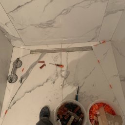 Luxs - Kompleksowe Remonty i Wykończenia - Układanie płytek imitujących marmur w kabinie prysznicowej, widoczny odpływ liniowy, przyssawka do płytek, poziomica, szpachelka, wiaderka z krzyżykami dystansowymi i młotek, w tle białe ściany...