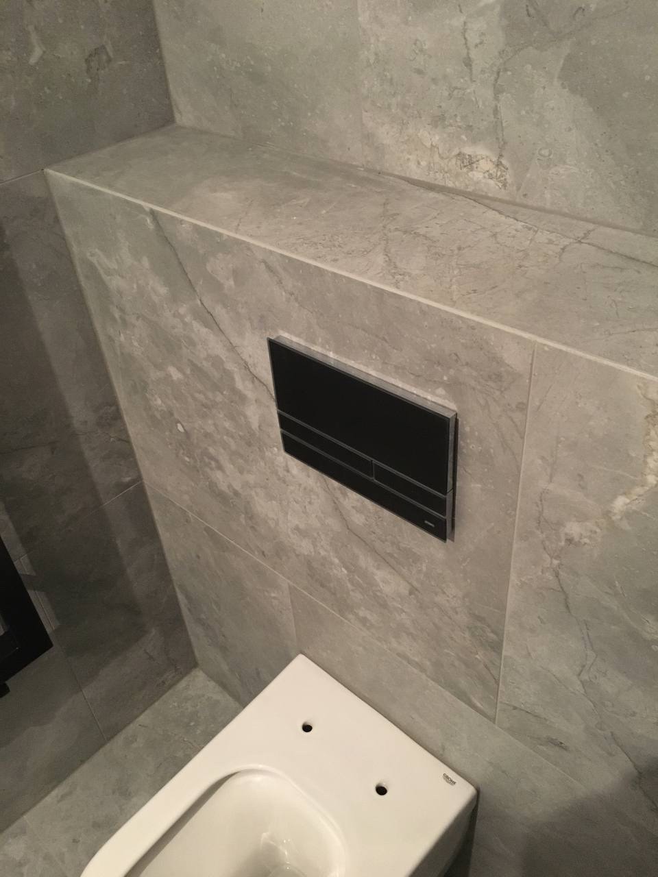 Świeżo wykończona łazienka z szarymi płytkami imitującymi marmur, wisząca toaleta marki Grohe i czarny panel sterujący spłuczką.