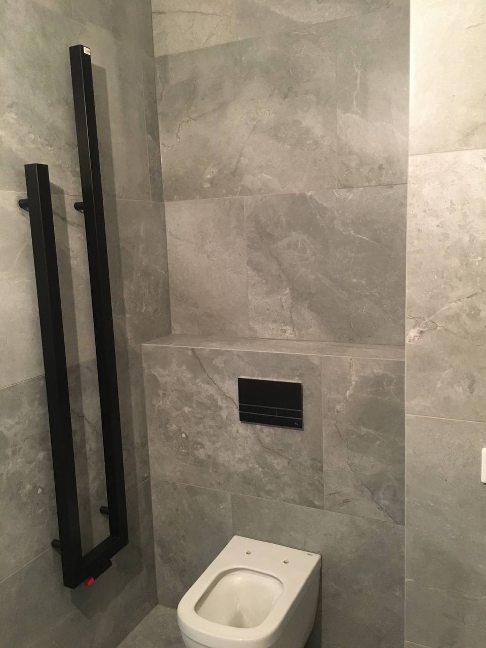 Wykończona łazienka z szarymi płytkami imitującymi marmur, wisząca toaleta z czarnym przyciskiem spłukującym oraz czarny, minimalistyczny grzejnik drabinkowy.