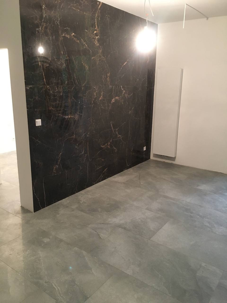 Wykończone wnętrze z dużymi, szarymi płytkami podłogowymi i ścianą wyłożoną czarnymi płytkami imitującymi marmur z wyraźnym użyleniem w odcieniach brązu i złota, widoczne gniazdko elektryczne...