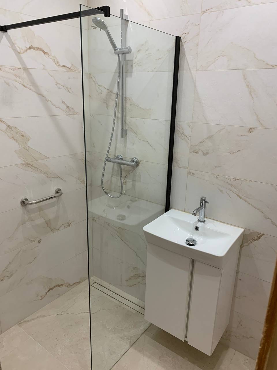 Wykończona łazienka w Warszawie: kabina prysznicowa ze szklaną ścianką i czarnym profilem, umywalka z szafką w kolorze białym, płytki imitujące marmur na ścianach i podłodze.