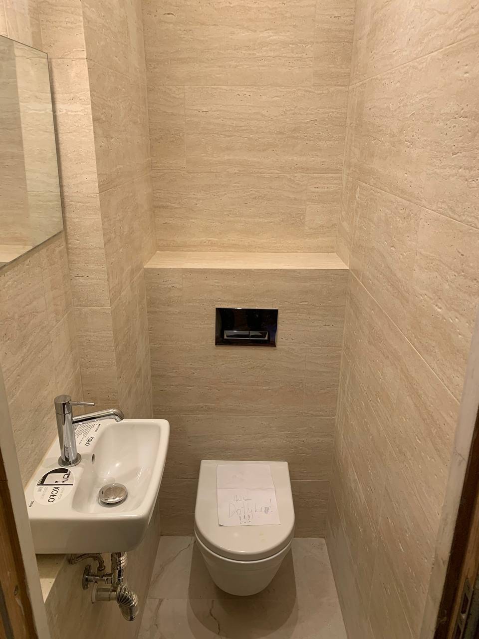 Wąska toaleta wykończona trawertynem z podwieszaną muszlą WC z kartką z napisem na desce oraz małą umywalką marki Koło, ze srebrną armaturą.