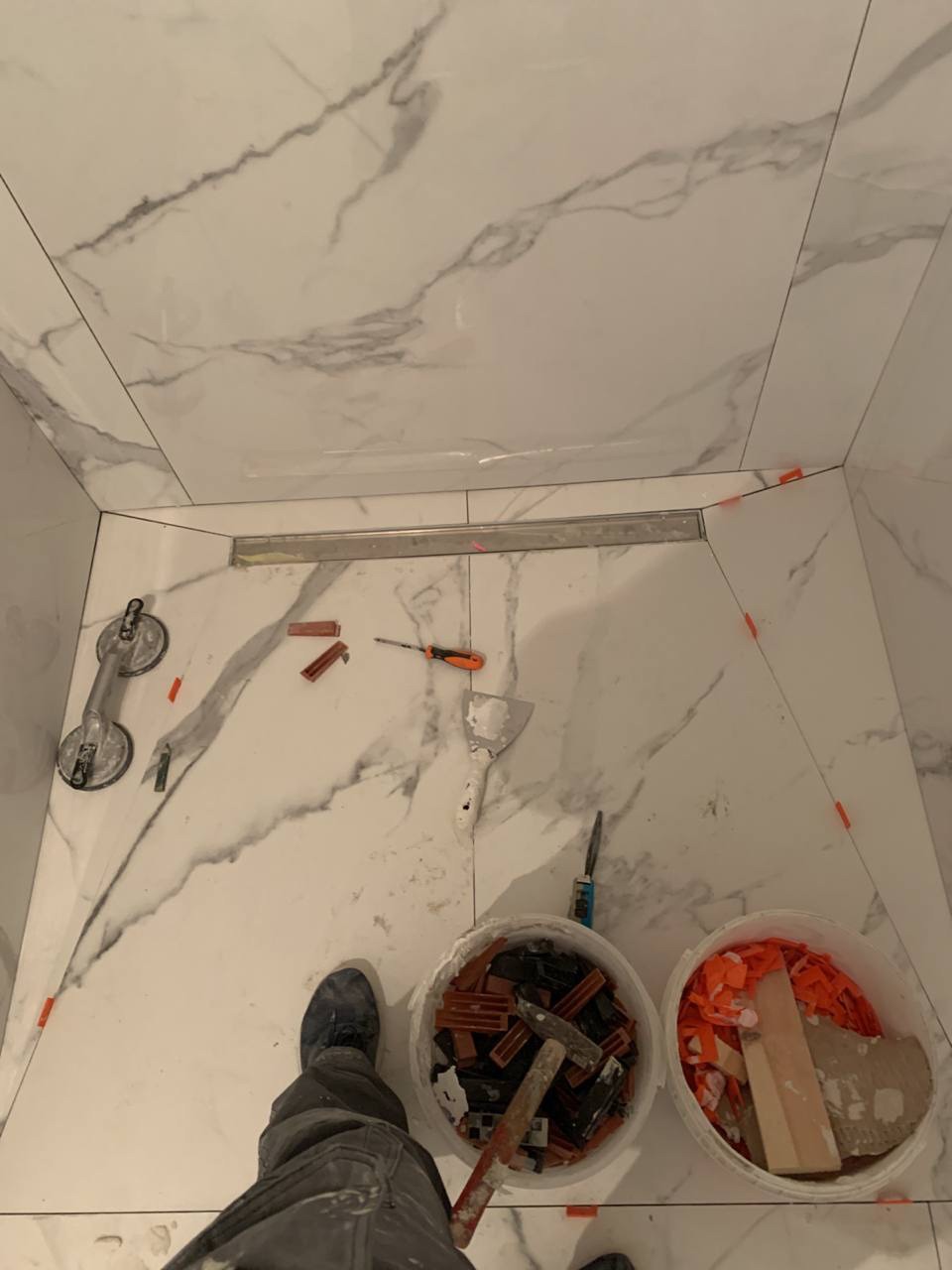 Układanie płytek imitujących marmur w kabinie prysznicowej, widoczny odpływ liniowy, przyssawka do płytek, poziomica, szpachelka, wiaderka z krzyżykami dystansowymi i młotek, w tle białe ściany...