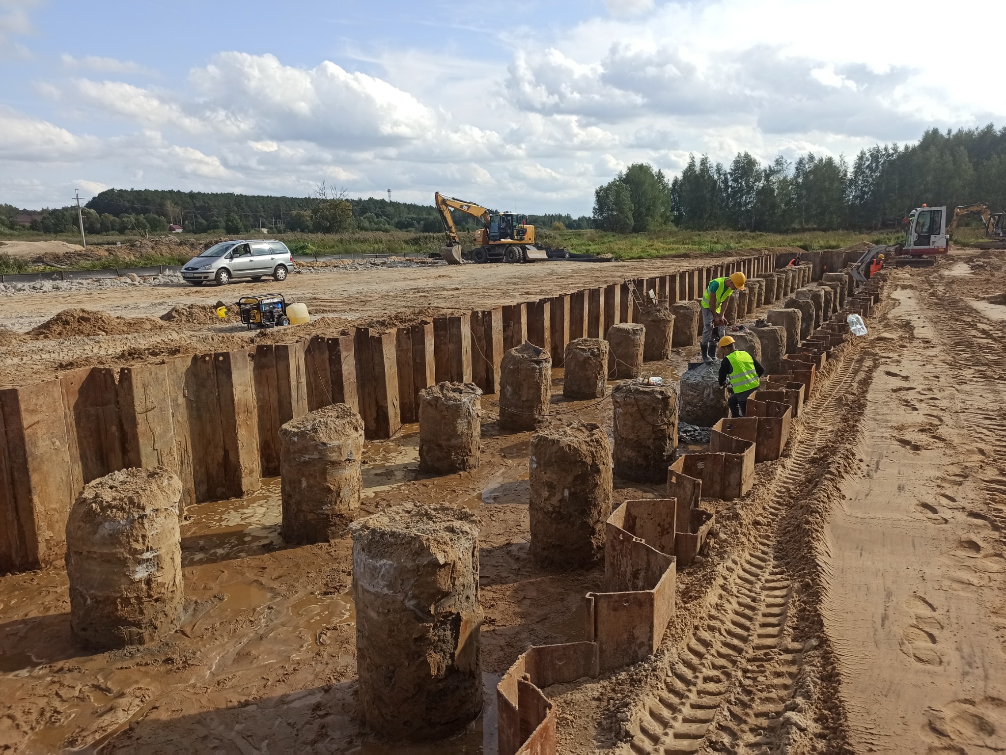 Budowa: Dwóch pracowników w żółtych kamizelkach i kaskach pracuje przy instalacji metalowych pali w wykopie, w tle widoczny jest samochód osobowy, koparka i spychacz, a także drzewa i niebo...