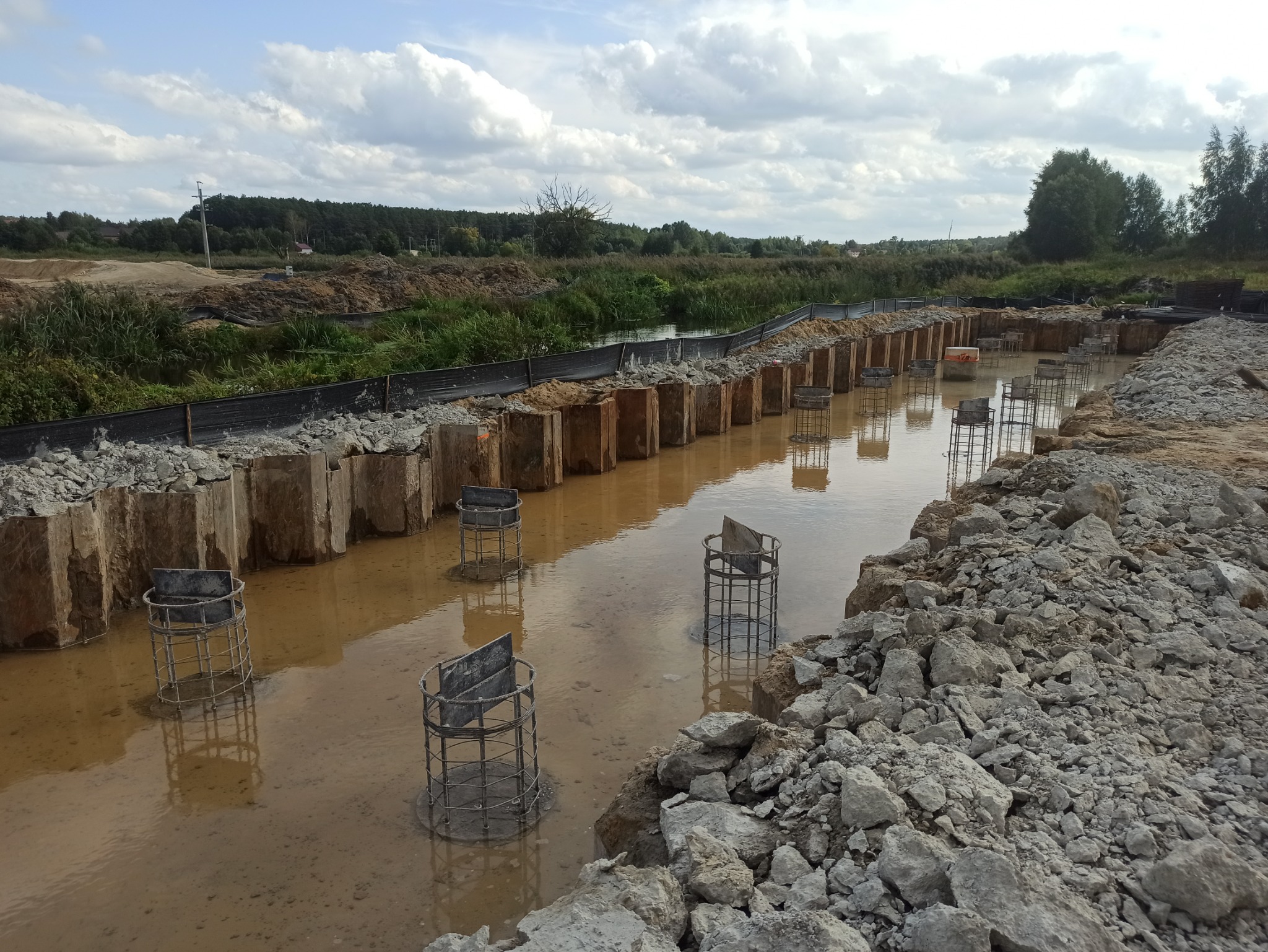 Budowa konstrukcji w wodzie: stalowe kosze zbrojeniowe osadzone w błotnistej wodzie, otoczone drewnianymi ścianami oporowymi i gruzem, krajobraz budowy z zielenią w tle.