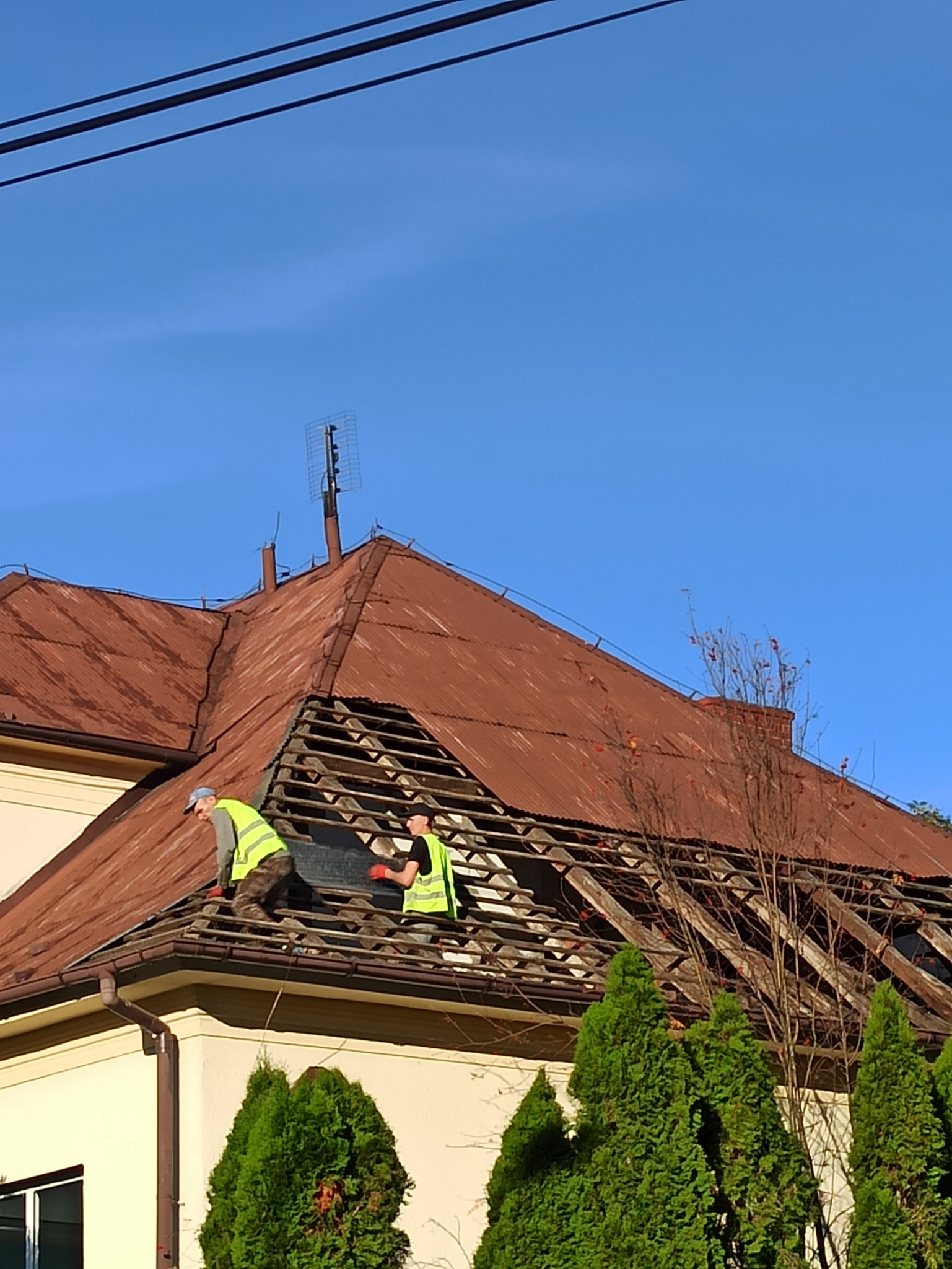 Dwaj robotnicy w jaskrawych kamizelkach pracują na dachu domu, wymieniając elementy konstrukcyjne pod starą, rdzawą blachą. Widoczny fragment elewacji budynku i zielone tuje w tle, na dachu antena...