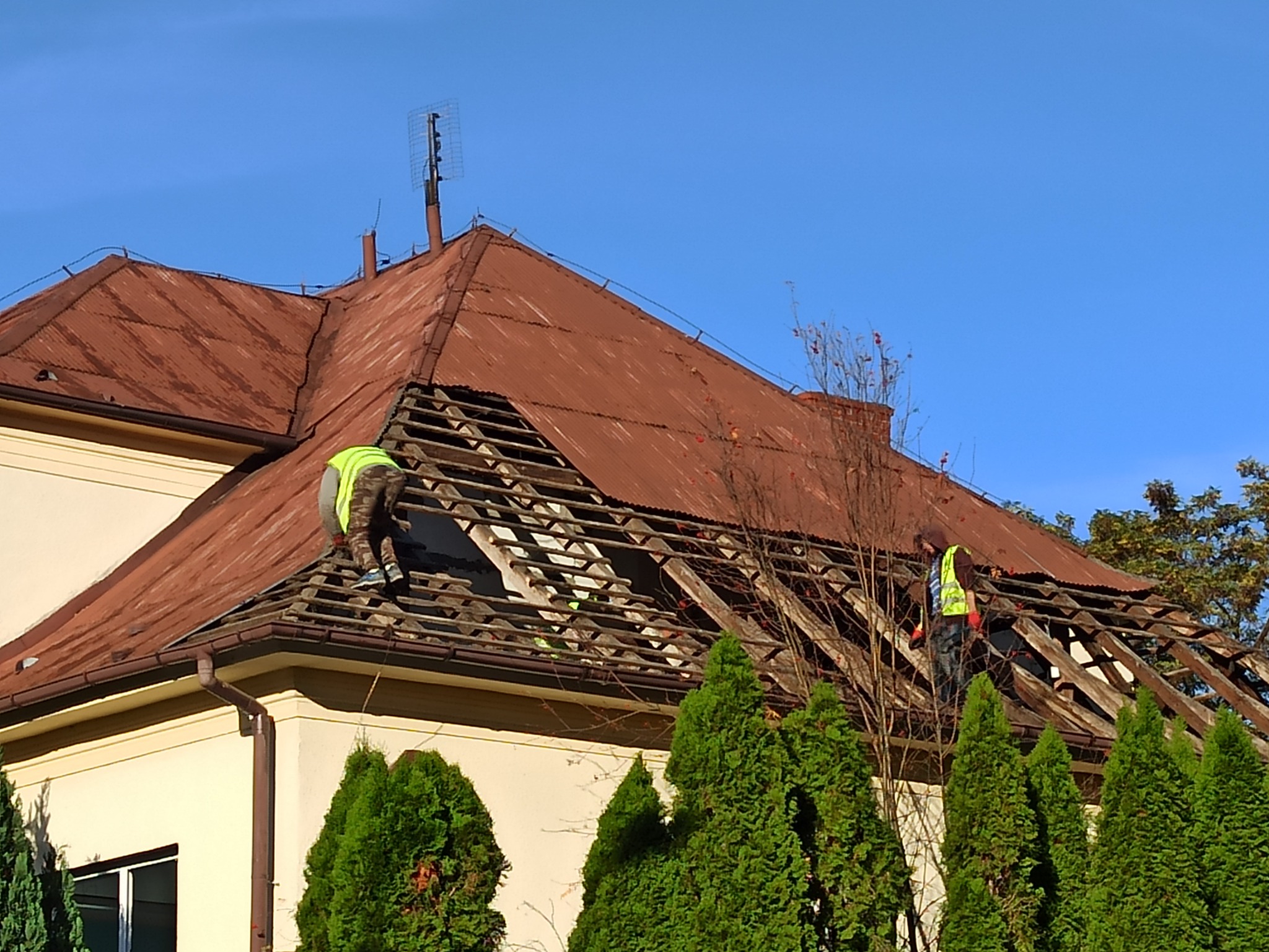 Demontaż starego, pordzewiałego dachu z blachy falistej na budynku jednorodzinnym, widoczni dwaj pracownicy w kamizelkach odblaskowych, fragmenty więźby dachowej odsłonięte, na pierwszym planie...