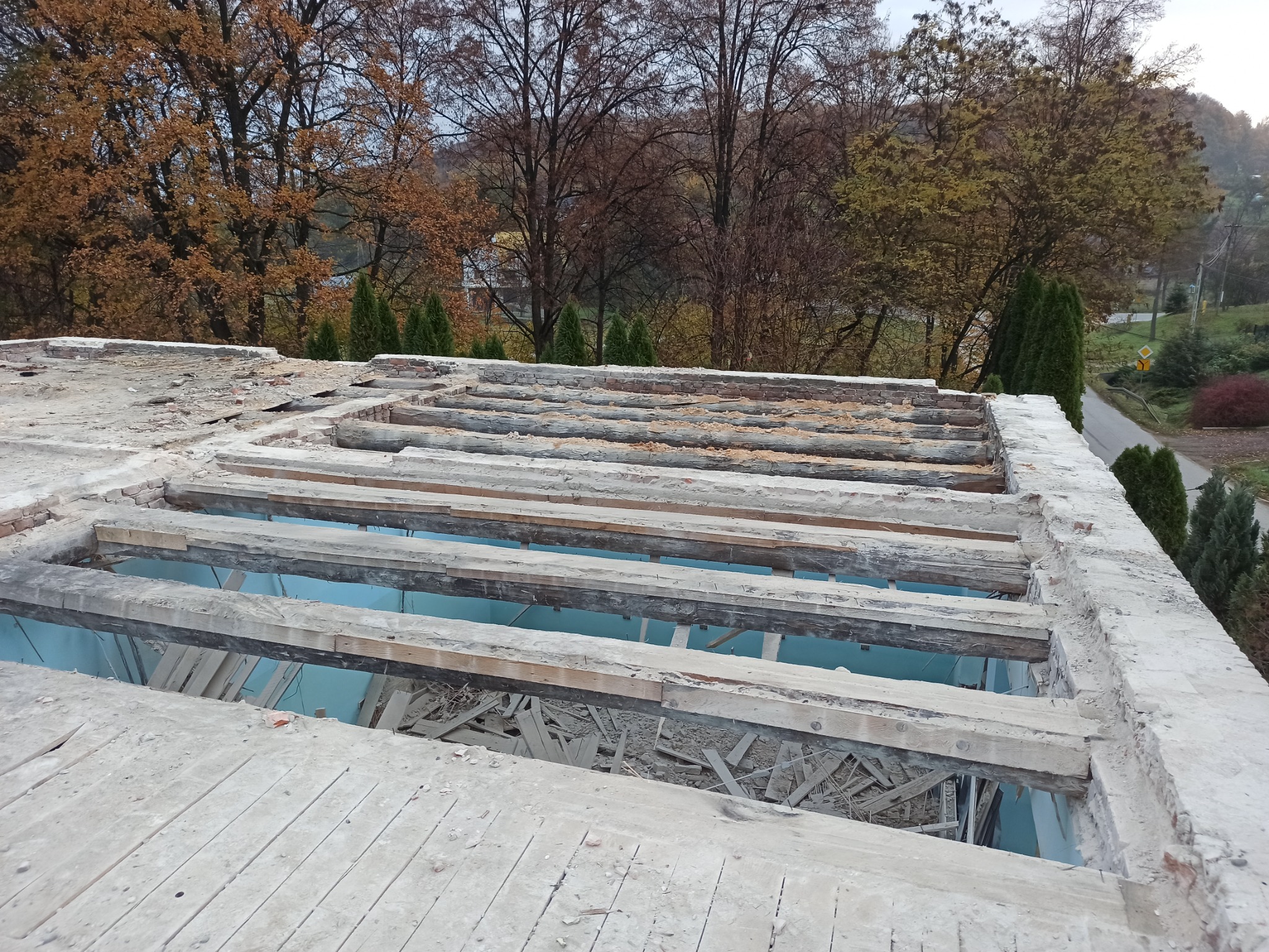 Widok z góry na częściowo zdemontowany dach budynku z odsłoniętymi drewnianymi belkami i fragmentami muru, w tle drzewa z jesiennymi liśćmi i droga.